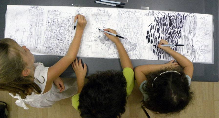 Three children drawing together on one long sheet of paper, seen from above