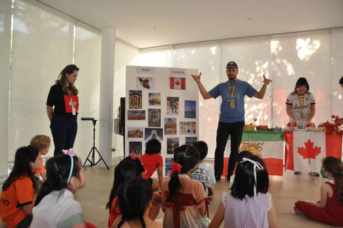 Children sitting together while parents present a country pavilion with flags and photographs