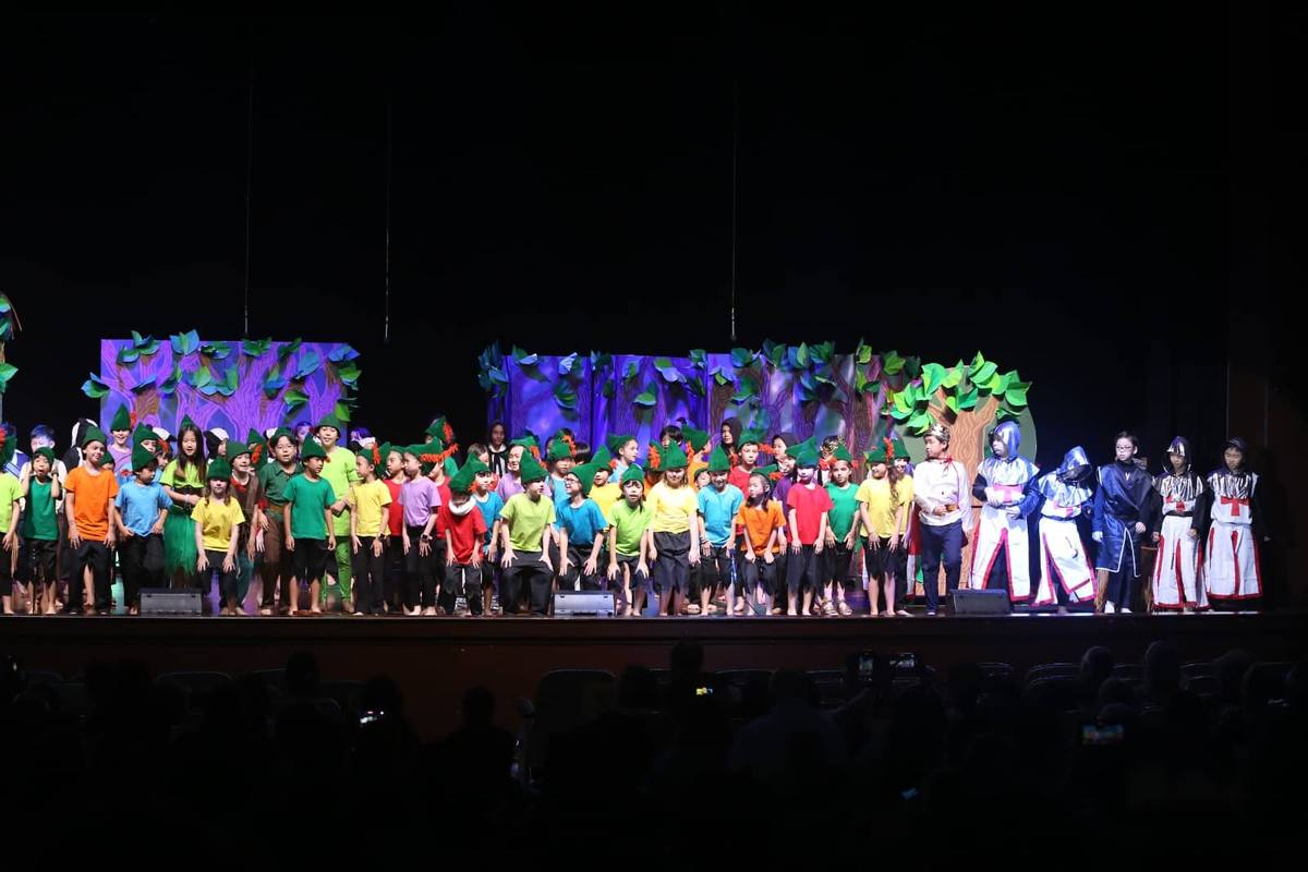 A full cast of children on a theatre stage in front of an audience, with forest scenery behind them