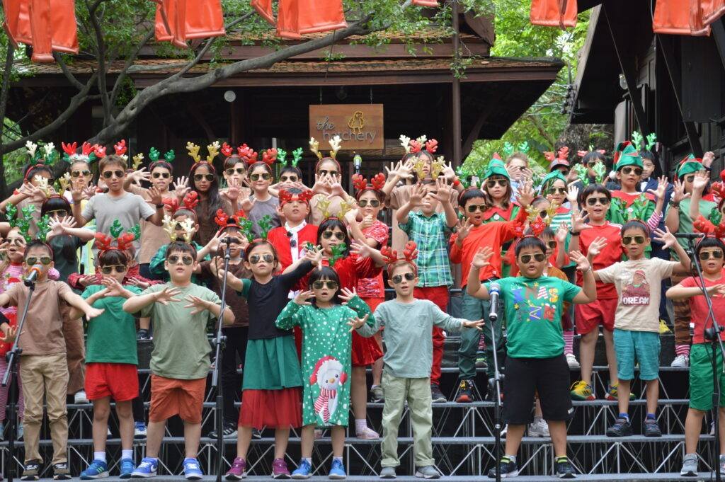 Children in festive costumes and antler headbands singing together on tiered staging