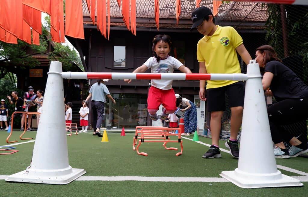 A young child jumps over a small hurdle on the field while an older child steadies the bar