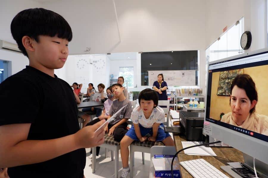 A child reads a question from a sheet of paper to a scientist on a large video call screen while classmates watch