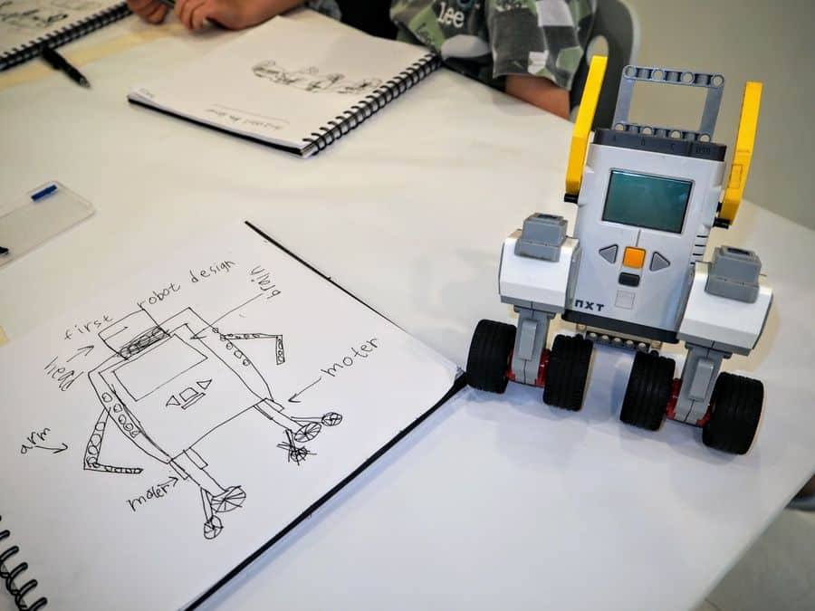 A small wheeled robot built from a kit stands on a table beside a child's labelled design sketch