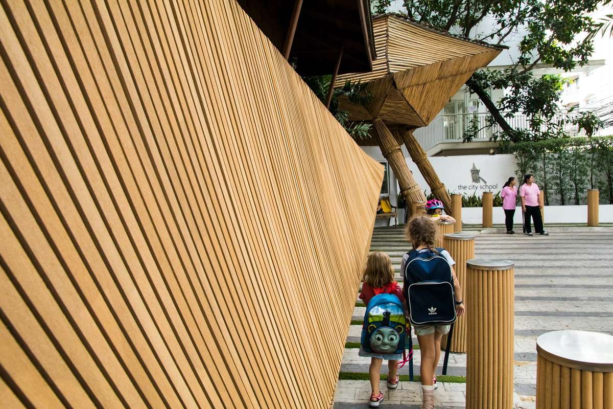 Two children with backpacks walking into the school entrance past its slatted bamboo wall