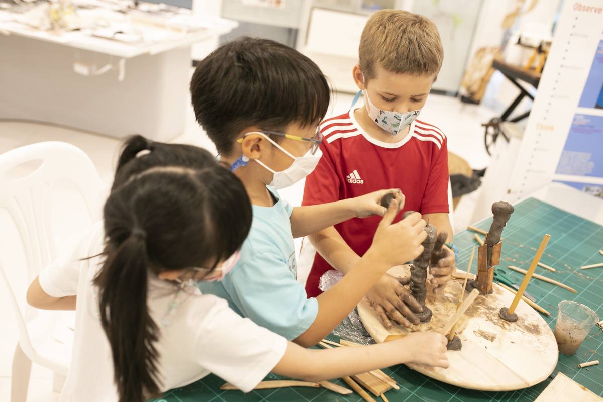 Three children shaping clay figures together on a wooden board in an atelier