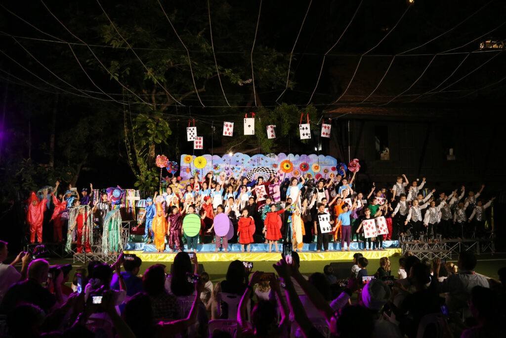 Children in costume on an outdoor stage at night while families watch and film from below